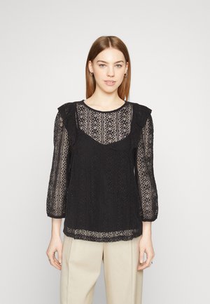 Μαύρη δαντελένια μπλούζα με υφασματάκια, κοντά φουσκωτά μανίκια και λαιμόκοψη με κυματισμούς. Συνδυασμένη με μπεζ παντελόνι.