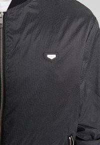 Svart bomberjacka med rutmönster, metallblixtlås och liten logotyp på bröstet. Slät textur och åtsittande design.