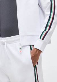 Άνδρας που φοράει λευκή φόρμα με ριγέ κορδέλα μάρκας σε πράσινο, λευκό και κόκκινο στο μανίκι και το πόδι του παντελονιού, με το χέρι στην τσέπη και σκούρο γκρι πουκάμισο από μέσα.