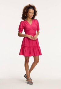 Lühike roosa kleit puffvarrukatega, vormitud büstiga ja kihiline seelik. Kandmist täiendab mustriliste sandaalide ja suurte rõngaskõrvarõngaste komplekt.