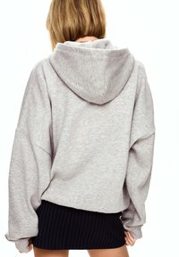 Šedá oversized mikina vyrobená z měkké tkaniny, s kapucí na šňůrku a spadlými rameny, kombinovaná s černou sukní s proužky.