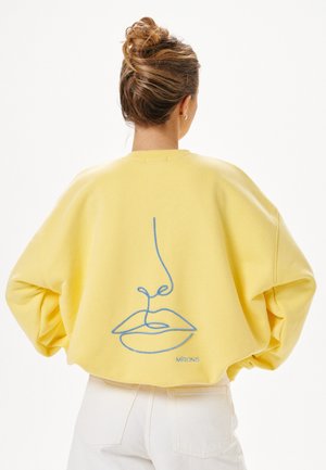 Woman with hair in a bun wearing a yellow sweater with a blue minimalist face line drawing on the back and white pants.