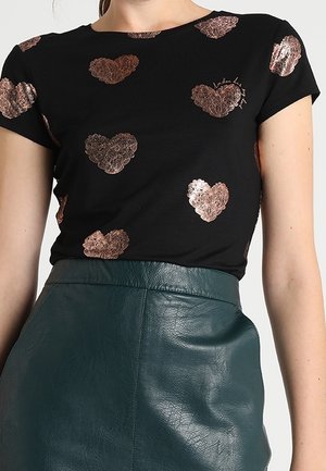 Top negro de manga corta con patrones metálicos de corazones en oro rosa, combinado con una falda de cuero verde oscuro de talle alto.
