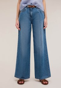 Jeans de mezclilla azul de pierna ancha con un lavado claro, que presentan una cintura alta y un diseño clásico de cinco bolsillos, acompañados de un cinturón decorativo.