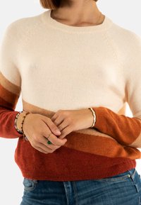Personne portant un pull coloré en crème, marron et rouille, un jean bleu, et plusieurs bagues et bracelets aux mains.