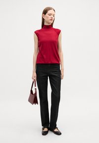Top senza maniche aderente rosso, design a collo alto; abbinato a pantaloni in denim neri, scarpe basse e una borsa bordeaux; stile minimalista.