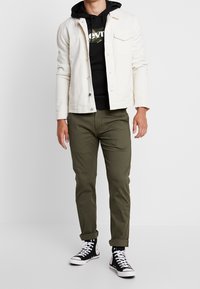 Giubbotto di denim bianco sopra un hoodie nero con un logo, pantaloni verde oliva e sneakers alte nere. Linee pulite e design casual.