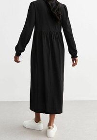 Schwarzes Maxikleid mit langen Ärmeln, strukturiertem Stoff, gerafftem Bund und elastischenÄrmelbündchen, kombiniert mit weißen Sneakers mit goldenen Akzenten.
