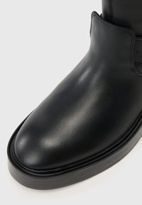 Μαύρη δερμάτινη μπότα μέχρι τον αστράγαλο με στρογγυλή μύτη, λεία υφή, ελάχιστες ραφές και επίπεδη μαύρη σόλα. Χωρίς ορατά υλικά ή σχέδια.