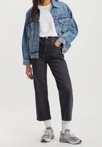 Chaqueta vaquera de color azul con dos bolsillos delanteros, llevada sobre una camiseta blanca. Vaqueros negros de tiro alto, zapatillas deportivas grises y calcetines blancos.