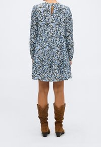 Femme portant une robe longue à manches longues bleue et blanche avec motifs floraux, une ouverture dans le dos et des bottes marron en daim montant jusqu'aux genoux, debout de dos.