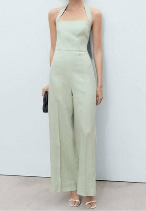 Donna che indossa una tuta verde chiaro a gambe larghe con scollo a halter, tiene una pochette nera e indossa sandali bianchi con punta aperta.