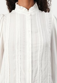 Vit blus med vertikala ränder, spetsdetaljer och en rufsig krage. Detaljer inkluderar knappar, ett lätt tyg och texturerade mönster.