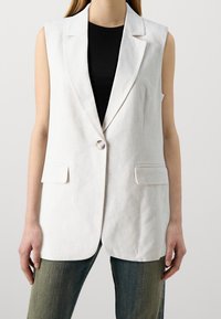 Bílý lněný bezrukávový blazer s jedním knoflíkem, dvěma předními kapsami a upraveným střihem, doplněný černým topem a modrými džíny.