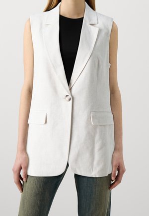 Blazer sans manches en lin blanc avec une fermeture à un bouton, deux poches avant et une coupe ajustée, associé à un haut noir et un jean bleu.