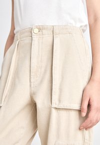 Beige Baumwoll-Cargohosen mit großen Seitentaschen, einem Knopfverschluss und einer lockeren Passform. Der Stoff hat eine weiche Textur und sichtbare Nähte.