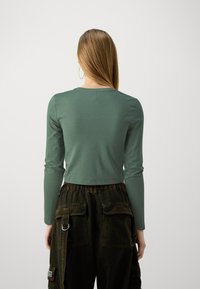 Langermet, cropped topp i dempet grønn stoff, med rund hals og glatt tekstur, kombinert med mørke cargobukser med lommer.