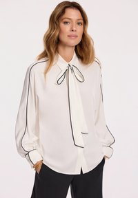 Biała bluzka z czarnymi lamówkami, z kołnierzykiem w kształcie kokardy, długimi rękawami, mankietami na guziki i gładką teksturą. Noszona z czarnymi spodniami.