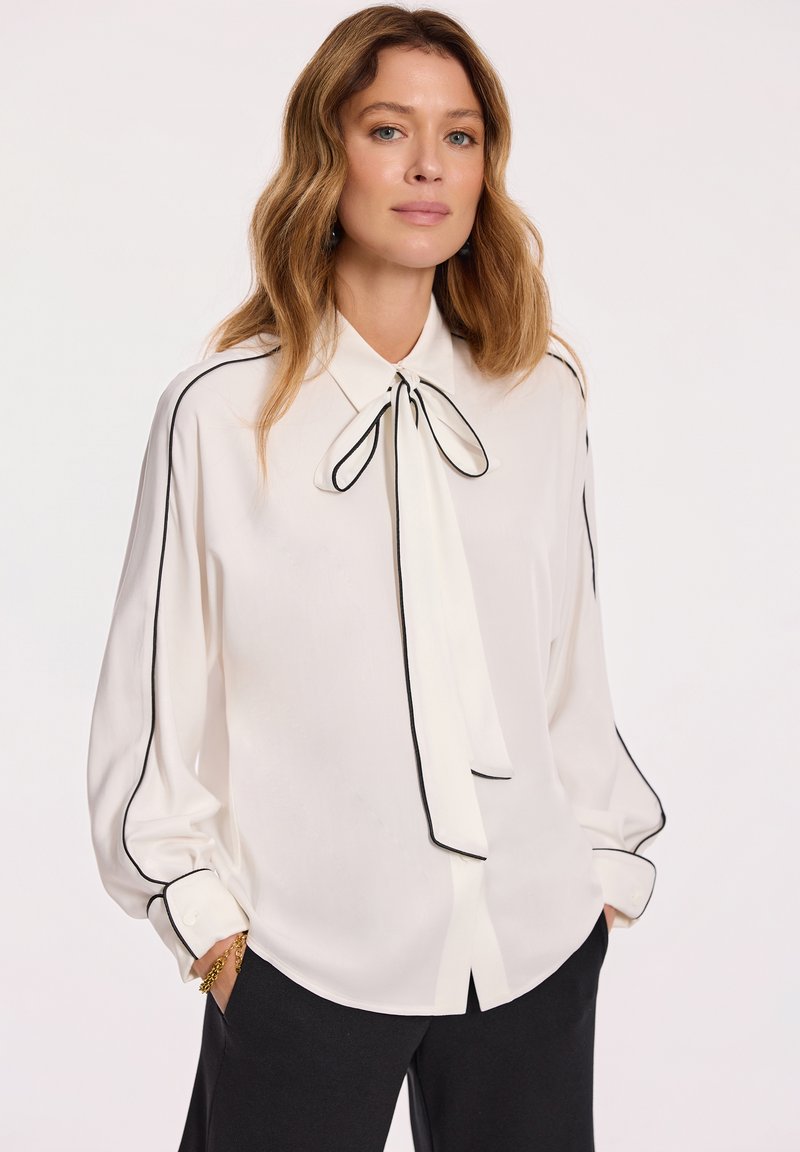 Biała bluzka z czarnymi lamówkami, z kołnierzykiem w kształcie kokardy, długimi rękawami, mankietami na guziki i gładką teksturą. Noszona z czarnymi spodniami.