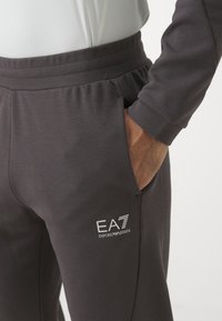 Pantaloni jogger grigi scuri con vita elasticizzata, tasche laterali e logo bianco EA7 Emporio Armani sulla coscia sinistra. Tessuto morbido.