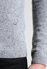 Maglione grigio lavorato a maglia con macchie nere, polsini e orlo a costine. Presenta un piccolo logo metallico vicino al fondo. Tessuto morbido e texturizzato.