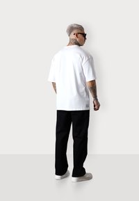 T-shirt en coton blanc à coupe décontractée, pantalon en denim noir et baskets blanches. Tatouages visibles sur les avant-bras et le cou, lunettes de soleil noires.