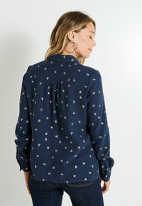 Camisa azul marino en tejido texturizado con patrones de lazo dorado, de manga larga y con detalle de espalda plisada. Apta para uso casual.