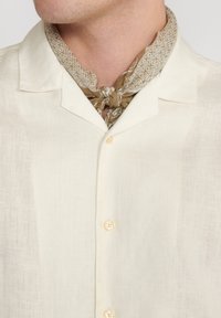 Camisa de lino beige claro con cuello tipo camp, adornada con una fila de botones claros. Un pañuelo marrón con estampado está atado al cuello.