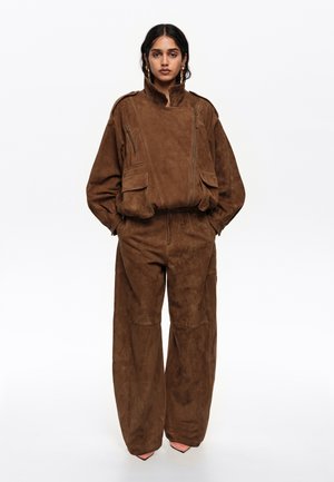 Mujer con chaqueta suelta de ante marrón con múltiples bolsillos y pantalones anchos a juego, de pie con las manos en los bolsillos contra un fondo blanco.