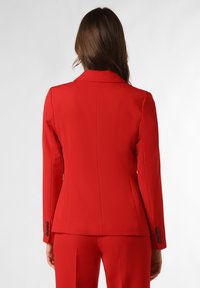 Blazer sur mesure rouge avec un col cranté, une fente unique et des accents de boutons sur les poignets. Tissu lisse avec une coupe structurée.