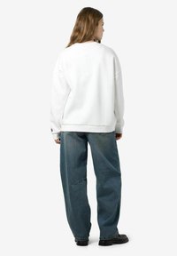 Witte oversized sweatshirt met verlaagde schouders, gecombineerd met losse donkerblauwe jeans, gedragen met zwarte schoenen, gezien vanaf de achterkant.