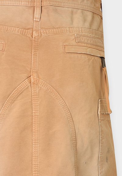 Κοντινό πλάνο καφέ παντελονιού εργασίας που δείχνει ενισχυμένες ραφές, πίσω τσέπες και ένα λουράκι με υφασμάτινη ετικέτα στην πλευρά.