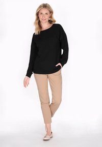 Fekete, hosszú ujjú felső, kerek nyakkivágással és elülső zsebbel, bézs, slim-fit nadrággal és világos színű lapos cipővel párosítva, fehér háttér előtt.