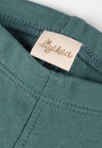 Nahaufnahme eines türkisfarbenen Hemdkragens, der die Nähte und ein gewebtes Etikett mit dem auffällig gestalteten Schriftzug "sigikid" zeigt.