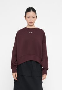 Κόκκινη μπλούζα με κοντό σχέδιο, με λευκό λογότυπο Nike στο στήθος. Μακριά μανίκια και μαλακή υφή. Σε συνδυασμό με μαύρη φούστα.