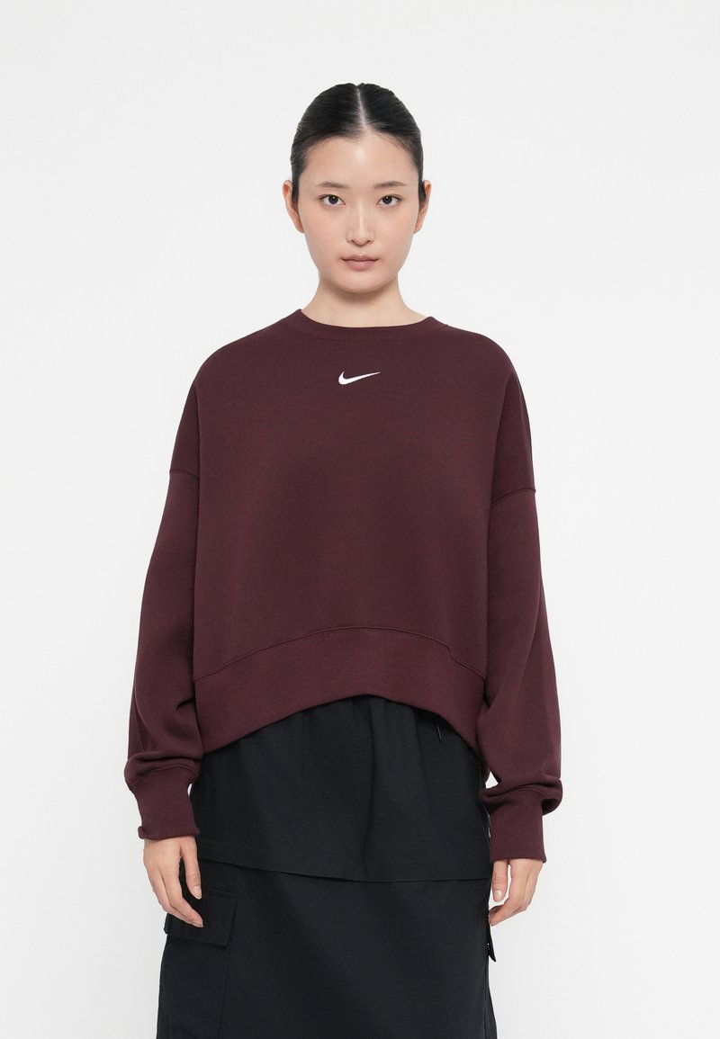 Κόκκινη μπλούζα με κοντό σχέδιο, με λευκό λογότυπο Nike στο στήθος. Μακριά μανίκια και μαλακή υφή. Σε συνδυασμό με μαύρη φούστα.