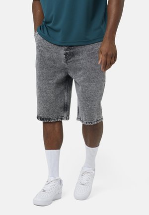 Man in grijze acid-wash denim shorts, witte sportsokken, witte sneakers en een teal shirt met handen in zijn zakken, staande op een witte achtergrond.