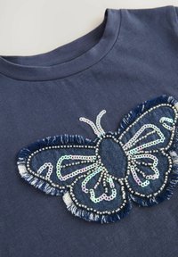 Marinblå t-shirt med en fjärilsdesign i paljetter och denim, med fransade kanter och metalliska detaljer. Rund hals.