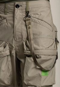 Beiga cargobyxor har flera fickor, en bältesöggla, ett avtagbart rem och en grön etikett på den nedre fickan. Strukturerad tygkvalitet och slät finish.