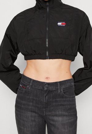 Gekürzte schwarze Reißverschlussjacke mit Tommy Jeans-Logo, kombiniert mit hoch taillierter dunkelgrauer Jeans, die den Bauchnabel freilegt, die Hände in die Hüften gestützt.
