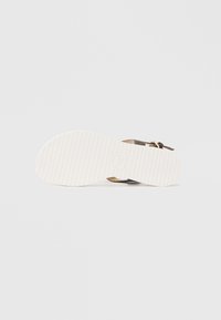 Hnědá sandála s texturovaným tmavým svrškem, plochou bílou gumovou podrážkou a minimalistickým designem s přezkou. Pohled ze spodní strany ukazuje žebrování.