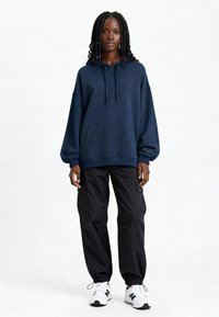 Tumši zils lielizmēra kapuce ar auklu, kangaru kabatu; apvienota ar melniem kravas biksēm un baltām sporta kurpēm ar melniem akcentiem.