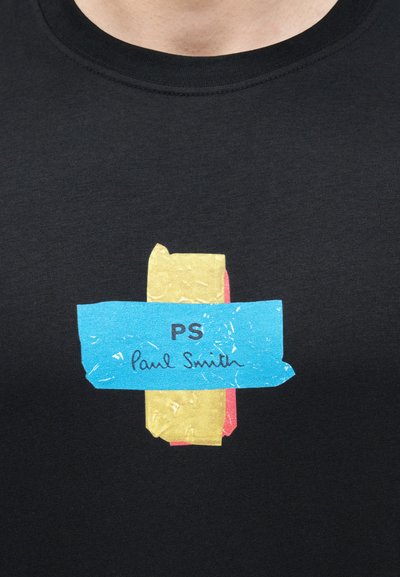 Chemise noire avec un graphisme de rubans jaune et rose croisés par un ruban bleu indiquant "PS Paul Smith" sur la poitrine.