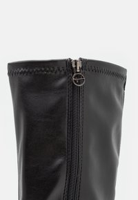 Bottes noires en cuir montant jusqu'au genou, avec une fermeture éclair latérale et des coutures décoratives le long du bord supérieur. Texture lisse avec une silhouette ajustée.