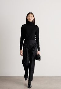 Γυναίκα που περπατά μπροστά φορώντας μαύρο ζιβάγκο, ψηλόμεσα μαύρα παντελόνια, μαύρες μυτερές μπότες και κρατώντας μια μικρή μαύρη τσάντα χειρός.