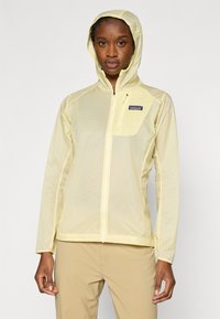 Ανοιχτό κίτρινο, ελαφρύ μπουφάν με φερμουάρ και κουκούλα, με υφή στην επιφάνεια και λογότυπο στο στήθος. Σχεδιασμένο για υπαίθρια χρήση.