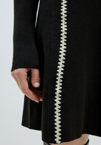 Schwarzes geripptes Kleid mit einem kontrastierenden weißen dekorativen Stich entlang der Seite. Nahaufnahme einer Hand, die auf dem Kleid platziert ist.