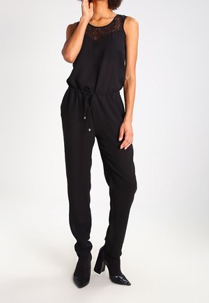 Kobieta ubrana w czarny kombinezon bez rękawów z koronkowym wykończeniem przy dekolcie i czarne buty na wysokim obcasie, stojąca na tle białego tła.