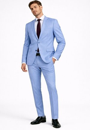 Homme debout en costume bleu clair, chemise blanche, cravate sombre, chaussures noires, une main dans la poche, sur fond blanc.