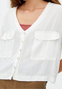 Blouse blanche à manches courtes avec un col en V, dotée de deux poches avant, de boutons perles et d'une texture légèrement translucide.